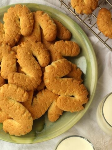 Sicilian S cookies piled on green plate with glasses of milk and cookies on wire rack around it.
