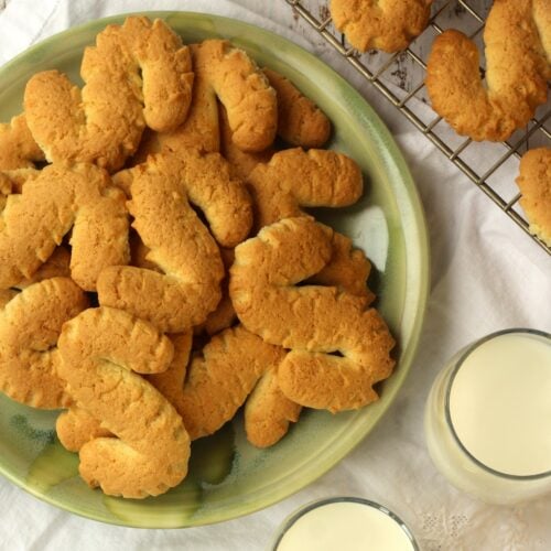 Sicilian S cookies piled on green plate with glasses of milk and cookies on wire rack around it.