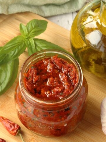 Sicilian sun dried tomato pesto (capuliatu) in jar next to garlic, chili peppers, basil leaves and olive oil bottle.