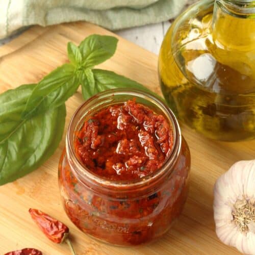 Sicilian sun dried tomato pesto (capuliatu) in jar next to garlic, chili peppers, basil leaves and olive oil bottle.