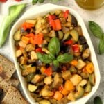 Summer vegetable stew in serving dish surrounded by bread, tomatoes, herbs and olive oil bottle.