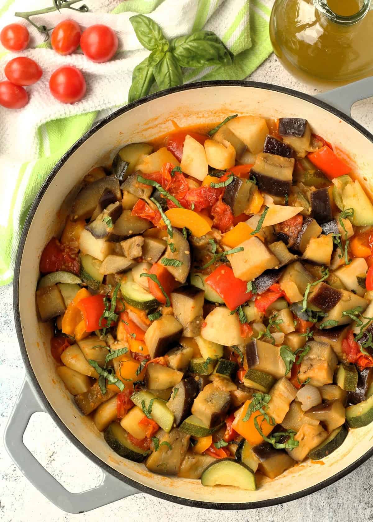 Sicilian summer vegetables stewed in skillet surrounded by basil leaves, cherry tomatoes and olive oil bottle.