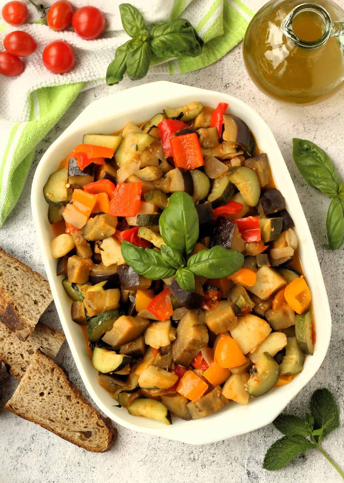 Summer vegetable medley (canazzo) with peppers, eggplant, zucchini and tomatoes in rectangular baking dish surrounded by olive oil and basil.