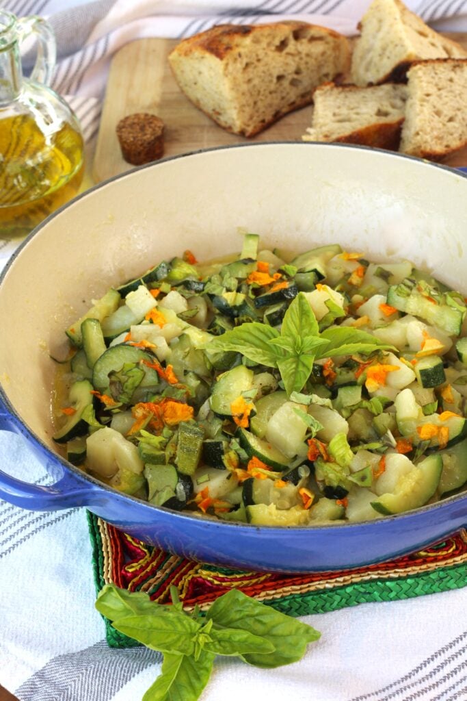 Sautéed zucchini flowers with zucchini and potatoes in skillet with olive oil and bread around it.