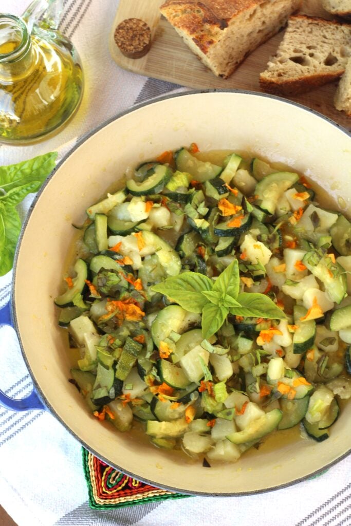 Large skillet filled with sautéed zucchini flowers, zucchini, potatoes and fresh basil with olive oil and bread around it.