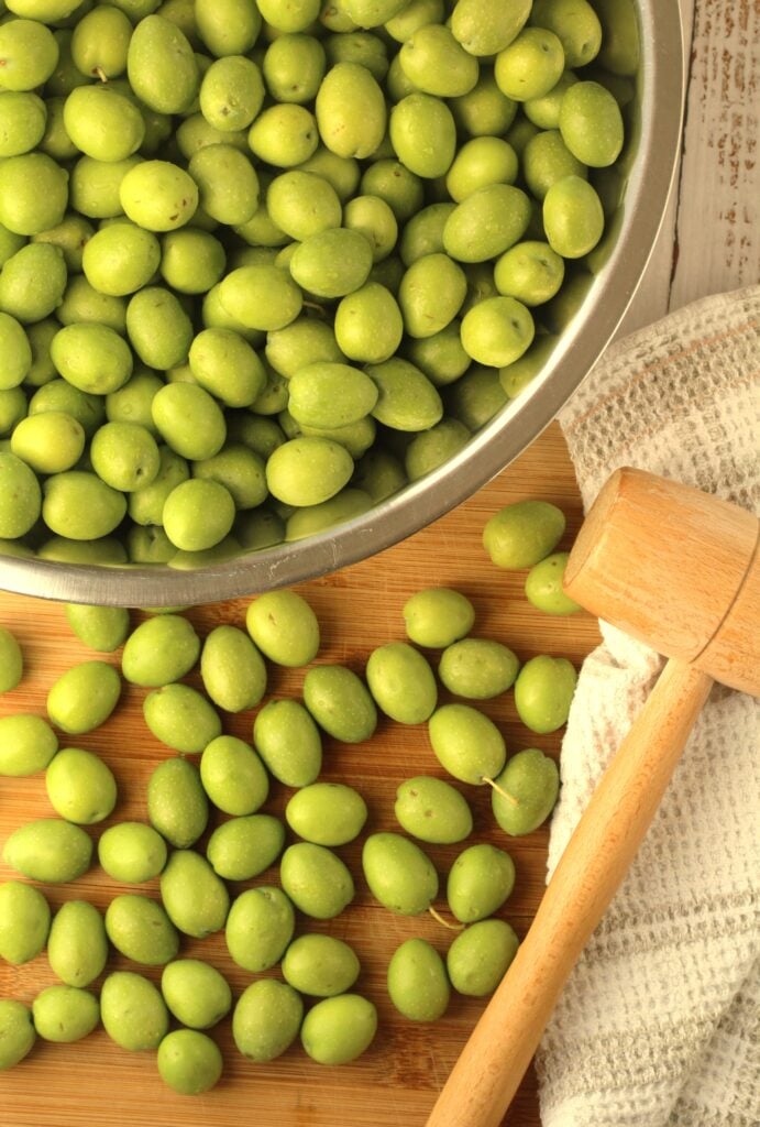 Bowl of green olives with several on a wood board next to meat mallet.