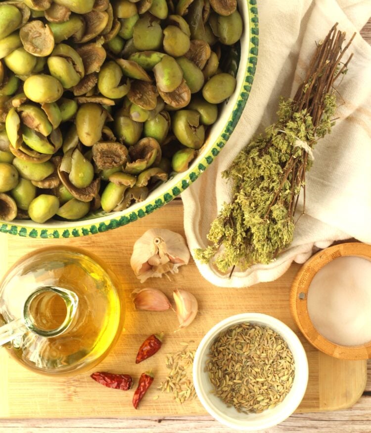 Cracked green olives in bowl surrounded by oregano, olive oil, salt, fennel seeds, garlic and pepperoncino.
