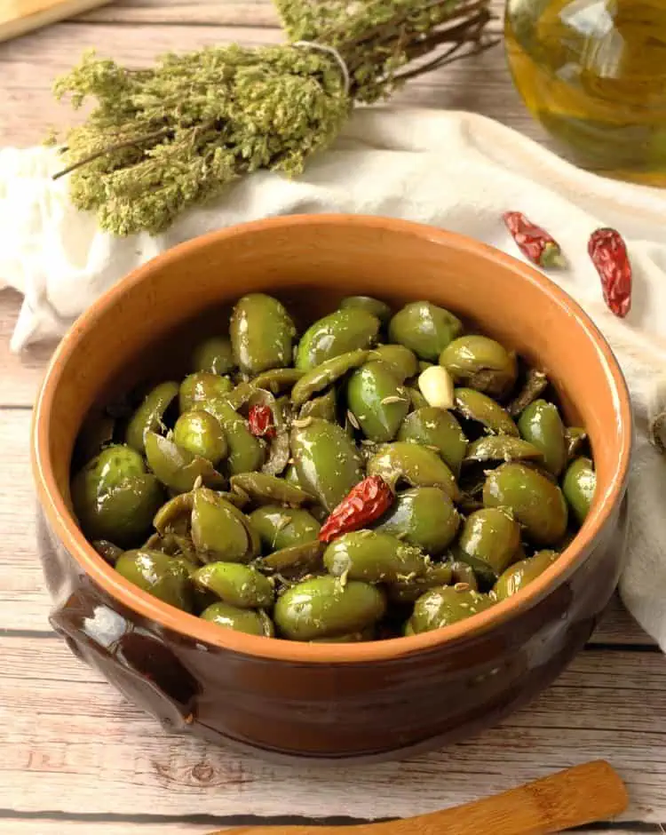 Sicilian cracked green olives seasoned in ceramic bowl surrounded by oregano branches and olives oil.