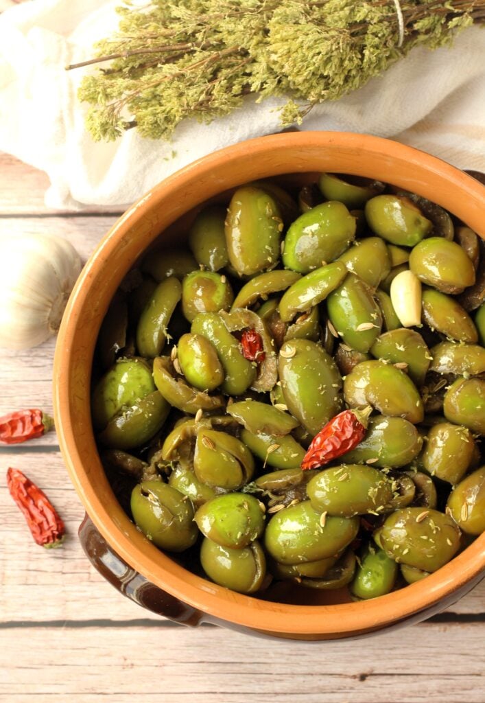 Cracked green olives seasoned in ceramic bowl with dry chilies and oregano branches around it.