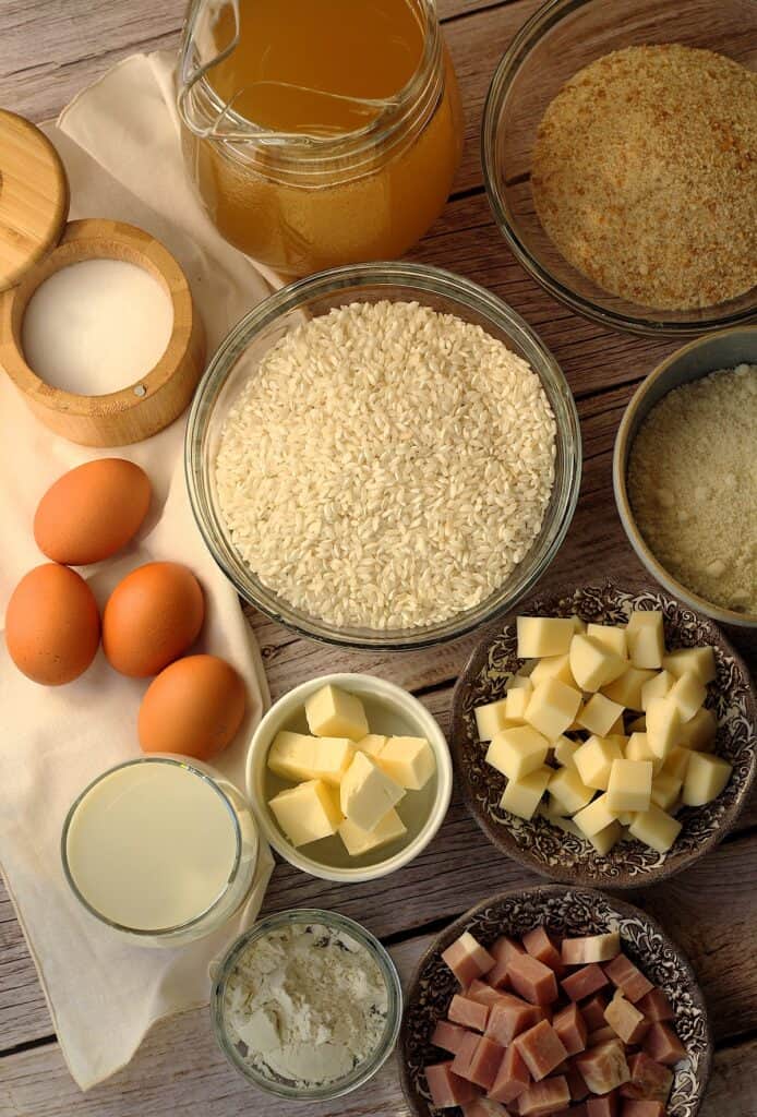 Bowls with rice, cheese, ham, butter, flour, salt, breadcrumbs, eggs and vegetable broth for making arancini.