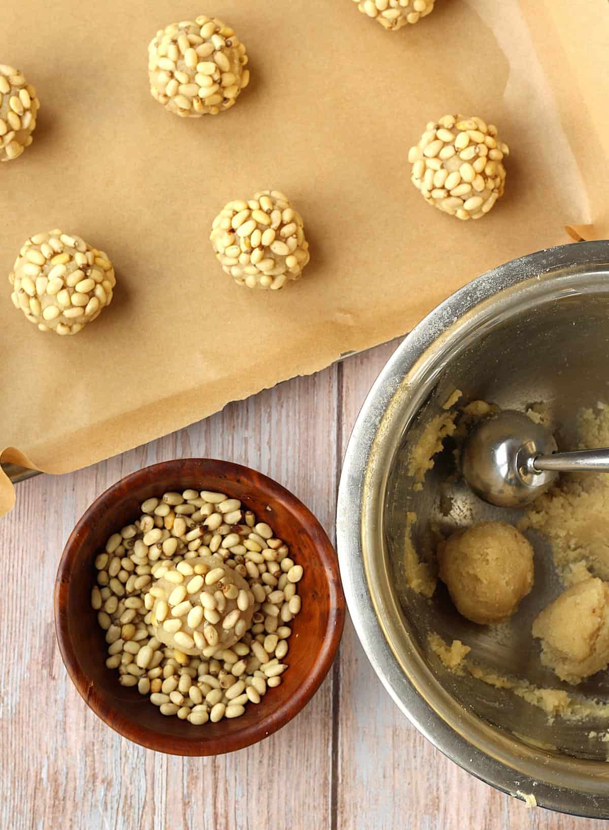 Scooped balls of almond dough rolled in bowl with pine nuts and placed on baking sheet.