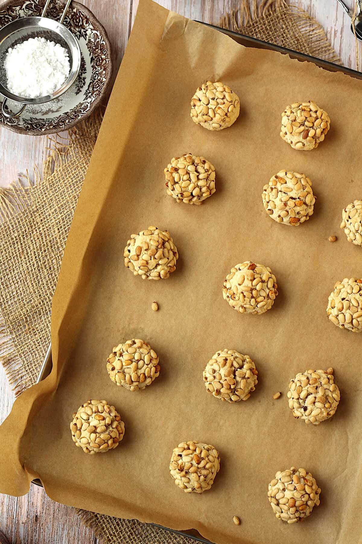 Baked pine nut cookies on baking sheet next to bowl of powdered sugar.