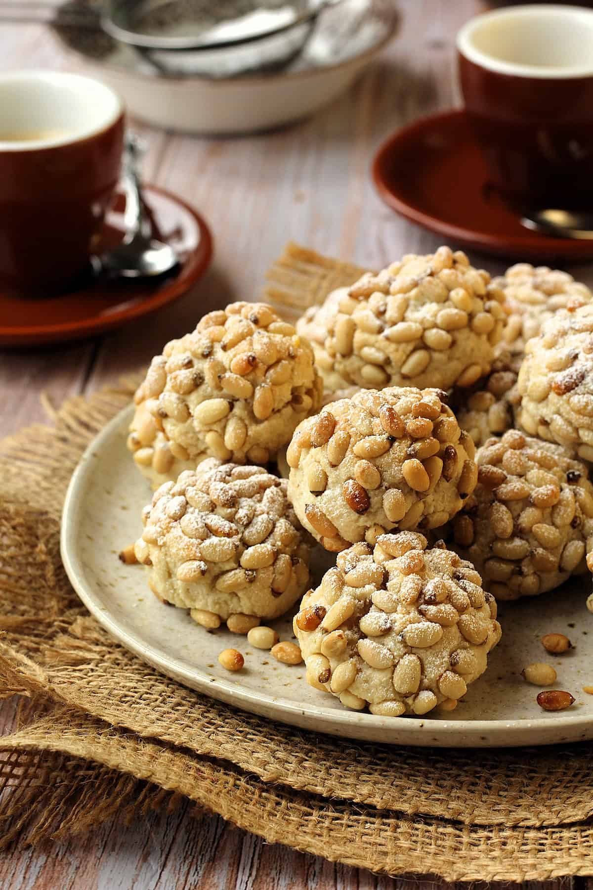 Round plate filled with pignoli cookies with coffee cups next to it.