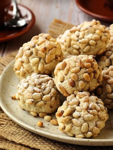 Pignoli cookies with pine nuts on round plate with coffee cups along side it.