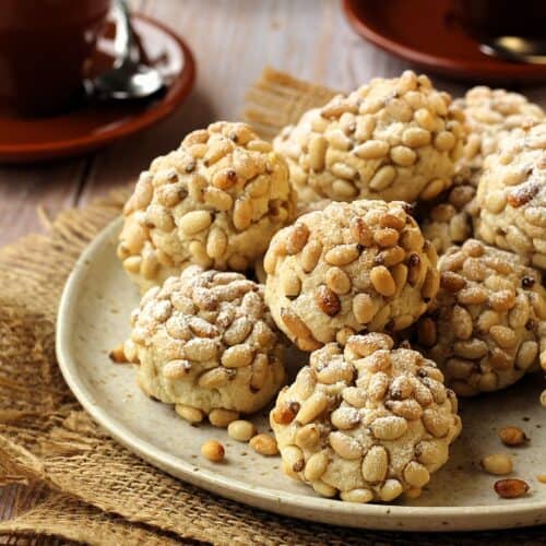 Pignoli cookies with pine nuts on round plate with coffee cups along side it.