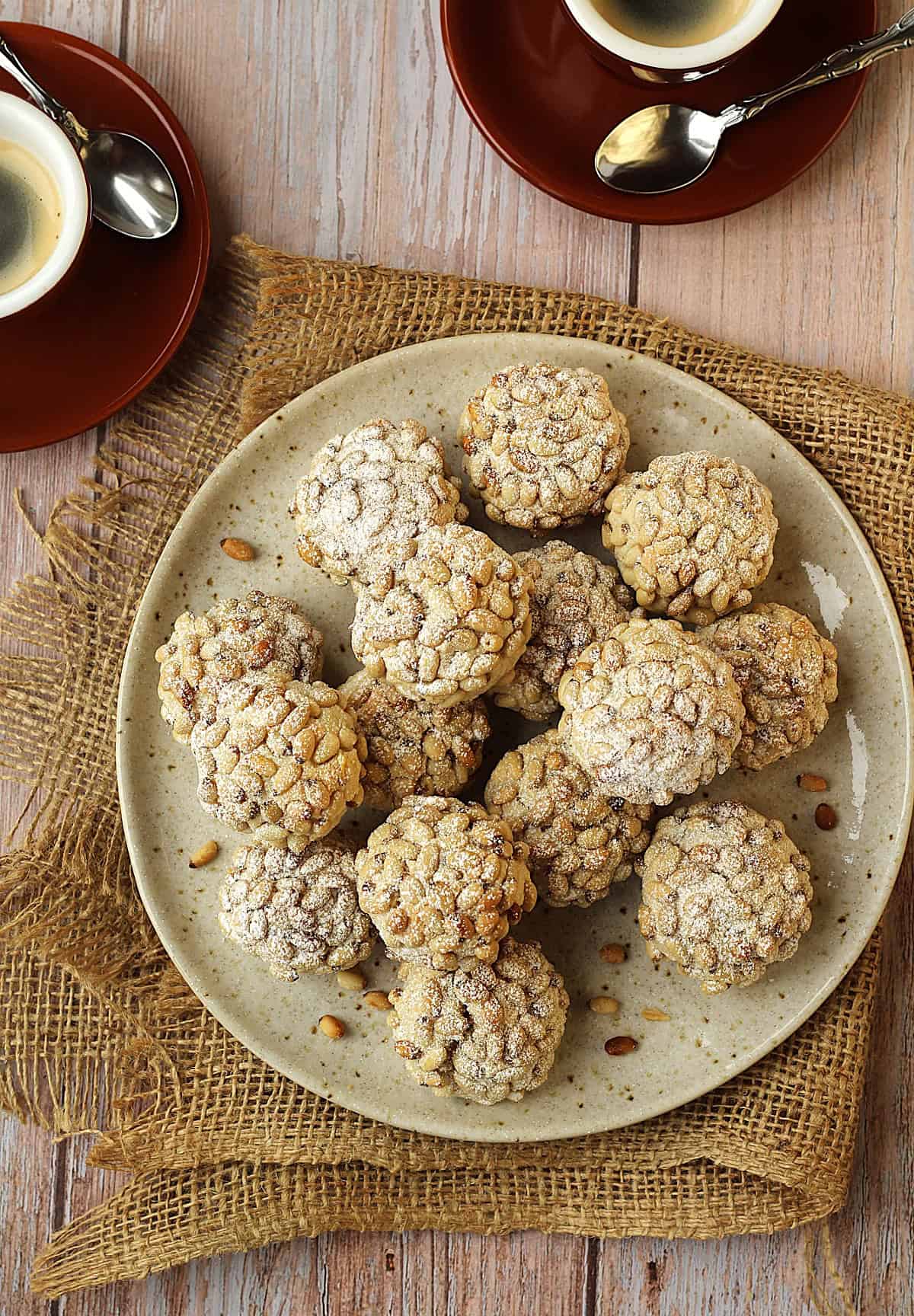 Plate filled with pignoli cookies (pine nut cookies) with cups of coffee along side it.