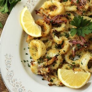 Baked breaded calamari and tentacles on oval white plate with lemon wedges and fresh parsley.