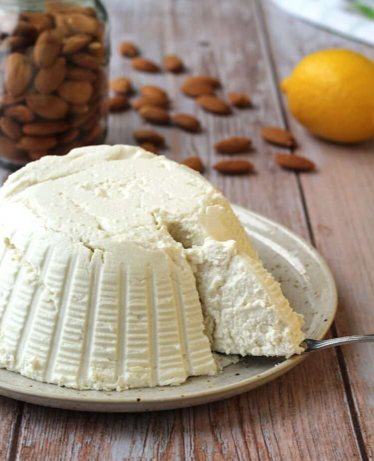 Vegan ricotta on plate with a spoon cutting into it with almonds and lemon behind it.