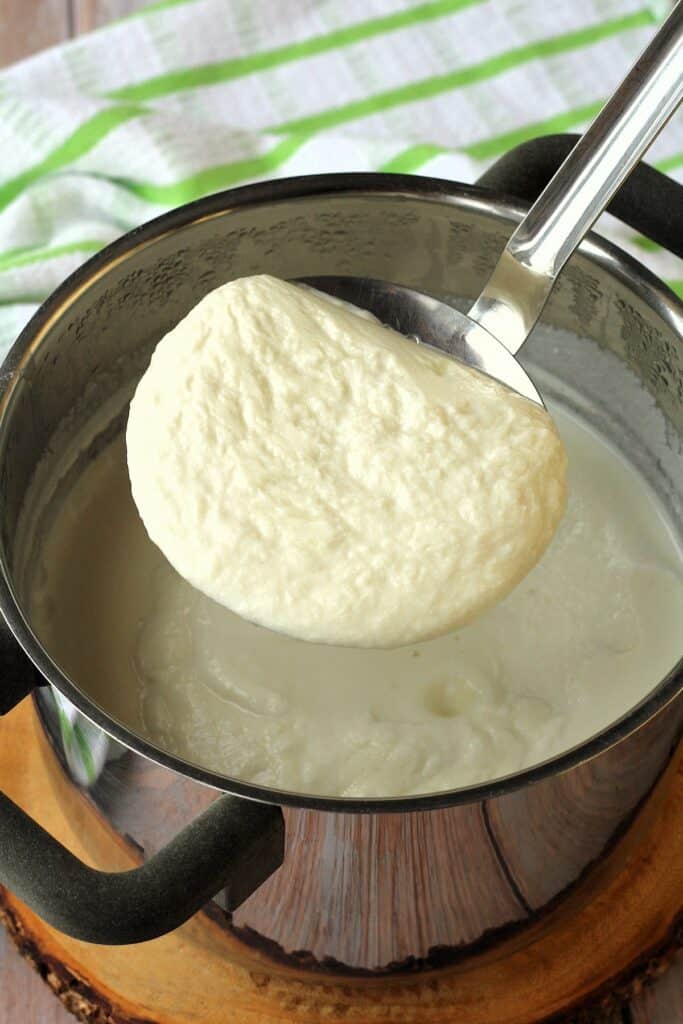 Slotted spoon holding curds from almond milk ricotta in sauce pan beneath it.