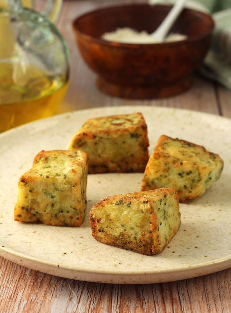 Fried frascatula squres on round plate with olive oil bottle and bowl of cheese behing it.