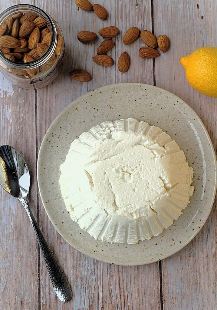 Overhead view of almond ricotta cheese on plate with spoon on side, jar of almonds and a lemon.