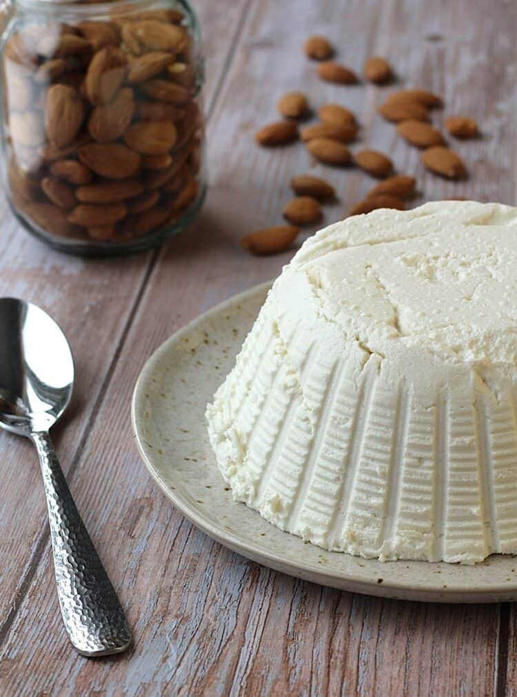 Almond milk ricotta on round plate with spoon on side and jar with almonds.