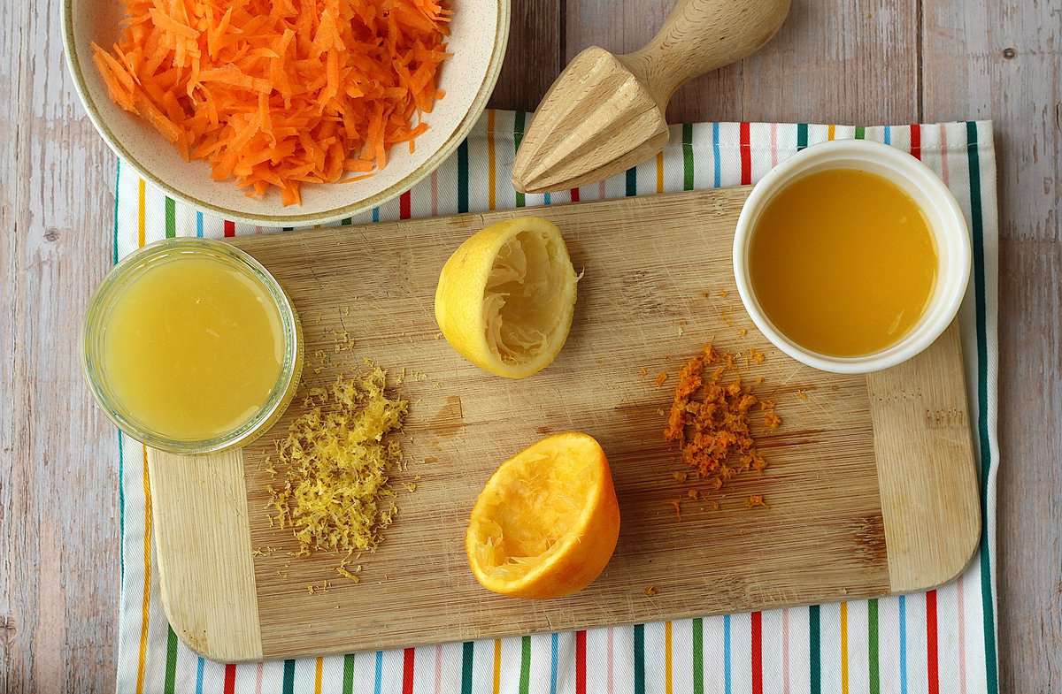 Bowl with grated carrots, zested and juiced orange and lemon on wood board.