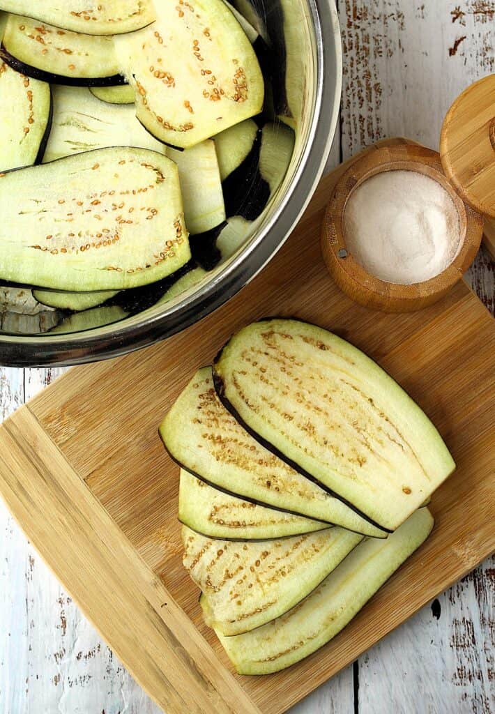 Sliced eggplant on wood board and in large bowl with salt.