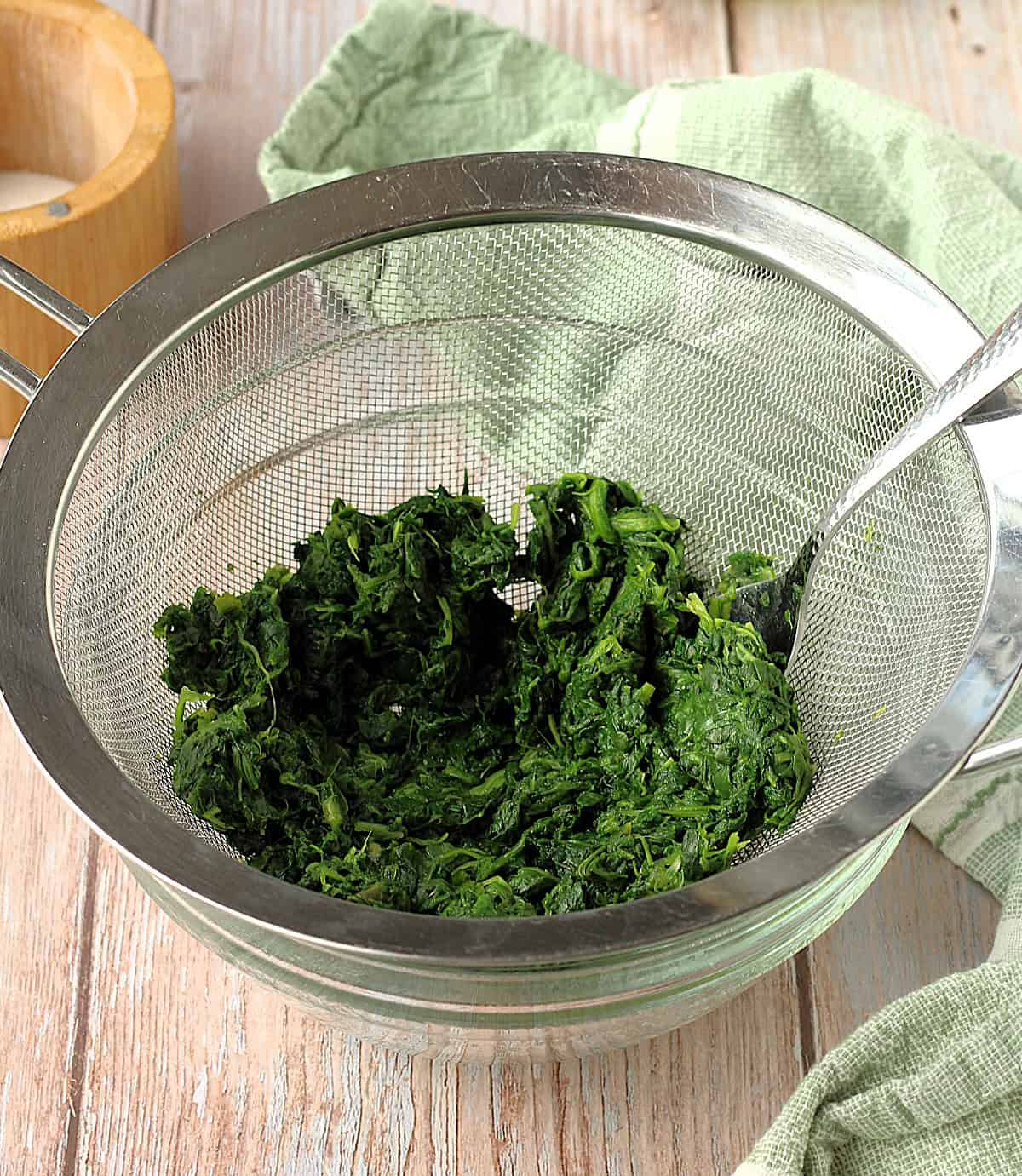 Drained spinach in fine mesh sieve over bowl with spoon.