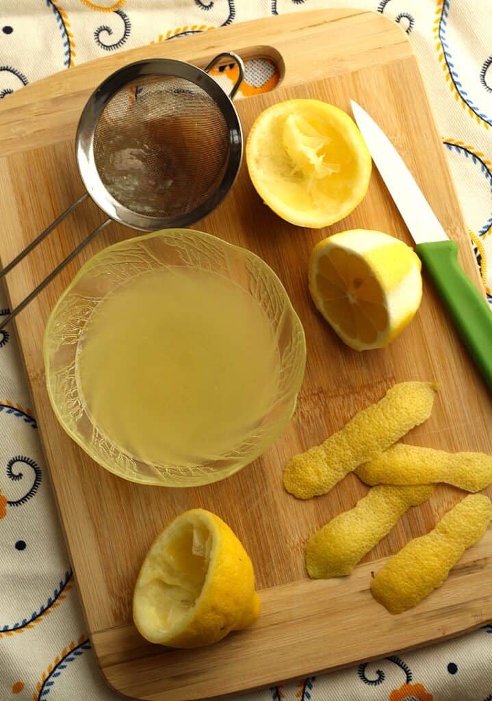 Lemon juice in bowl next to squeezed lemon halves and lemon rind strips on wood board.