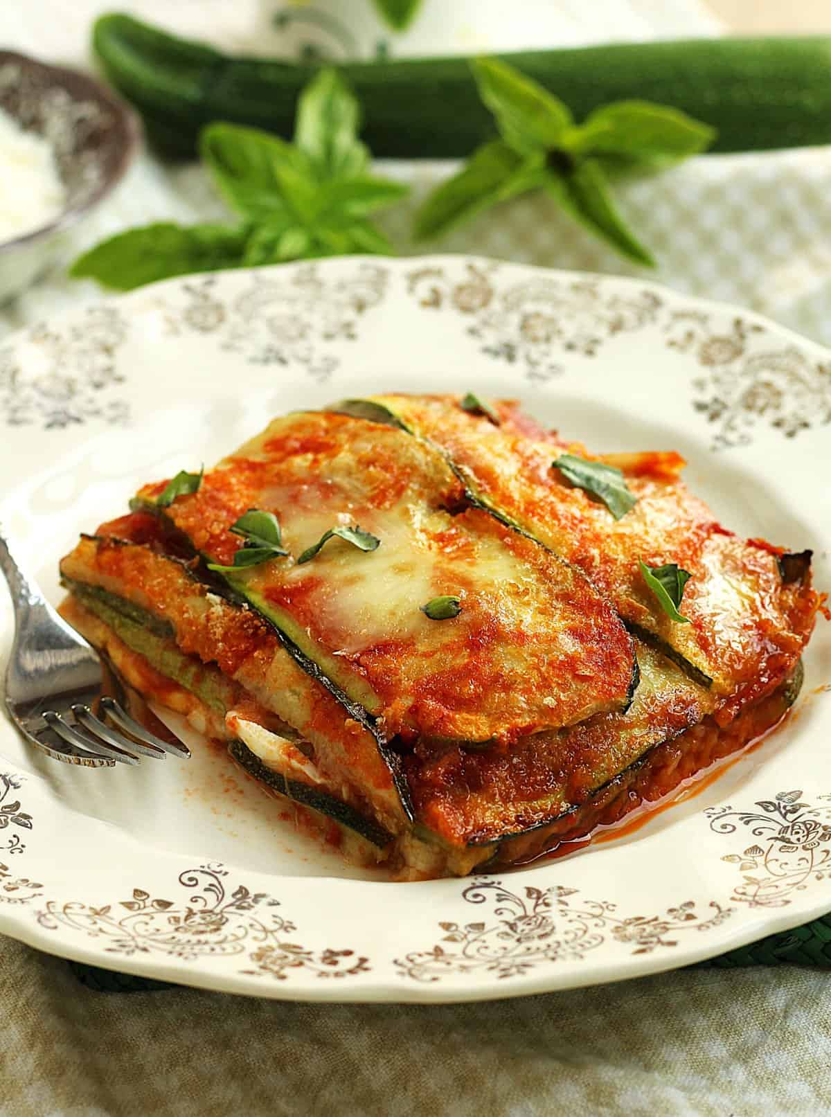 Square of zucchini parmesan on plate with fork topped with fresh basil.