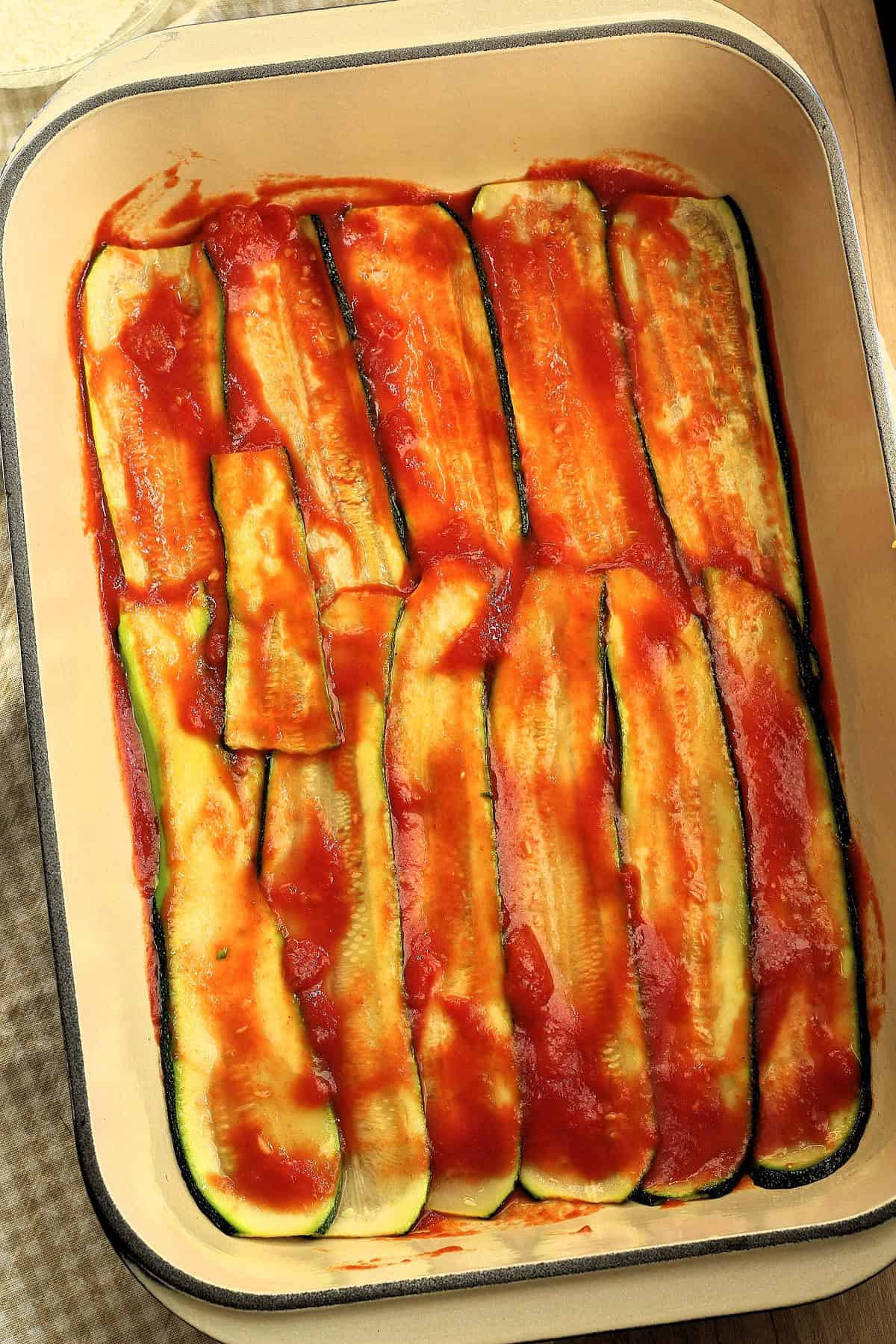 Sliced zucchini coated with a layer of tomato sauce in casserole dish.