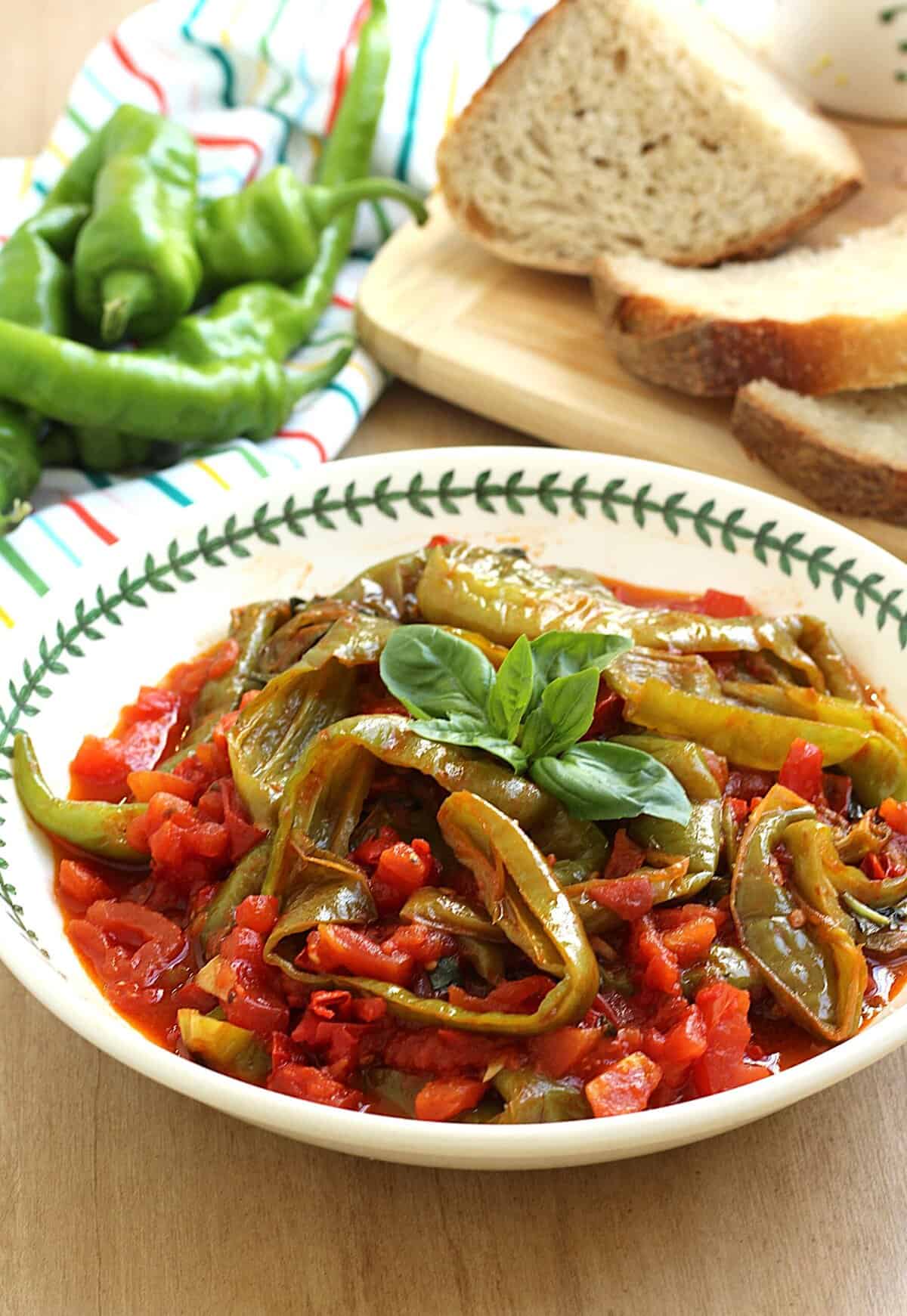 Pan-fried long peppers with tomatoes in bowl topped with basil and bread slices in background.