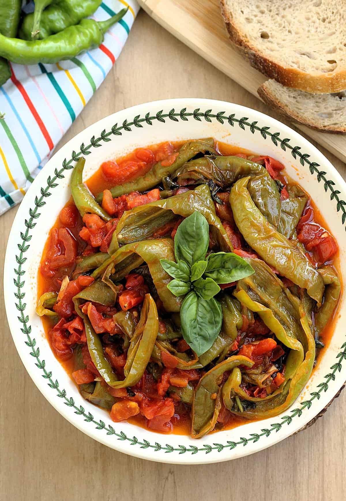 Pan-fried Italian friggitelli peppes with tomatoes in bowl topped with basil and bread on side.