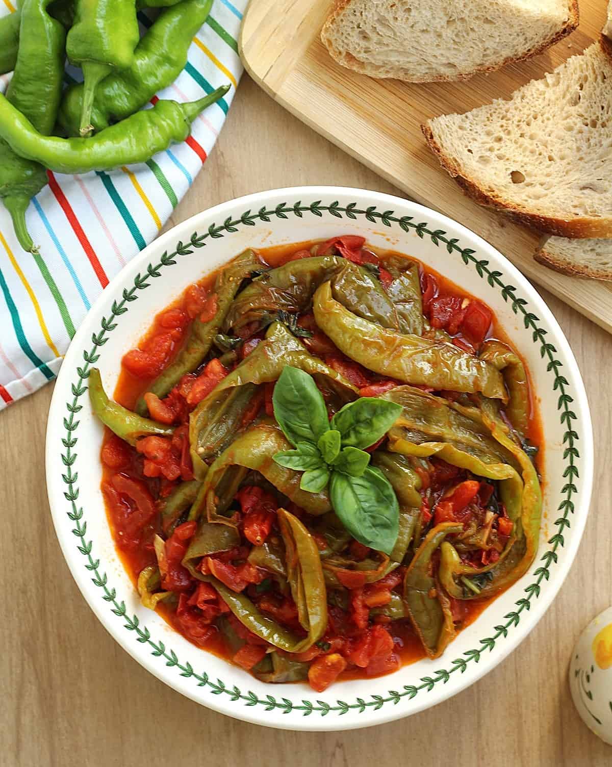 Pan-fried Italian peppers with tomatoes in bowl topped with basil next to bread.