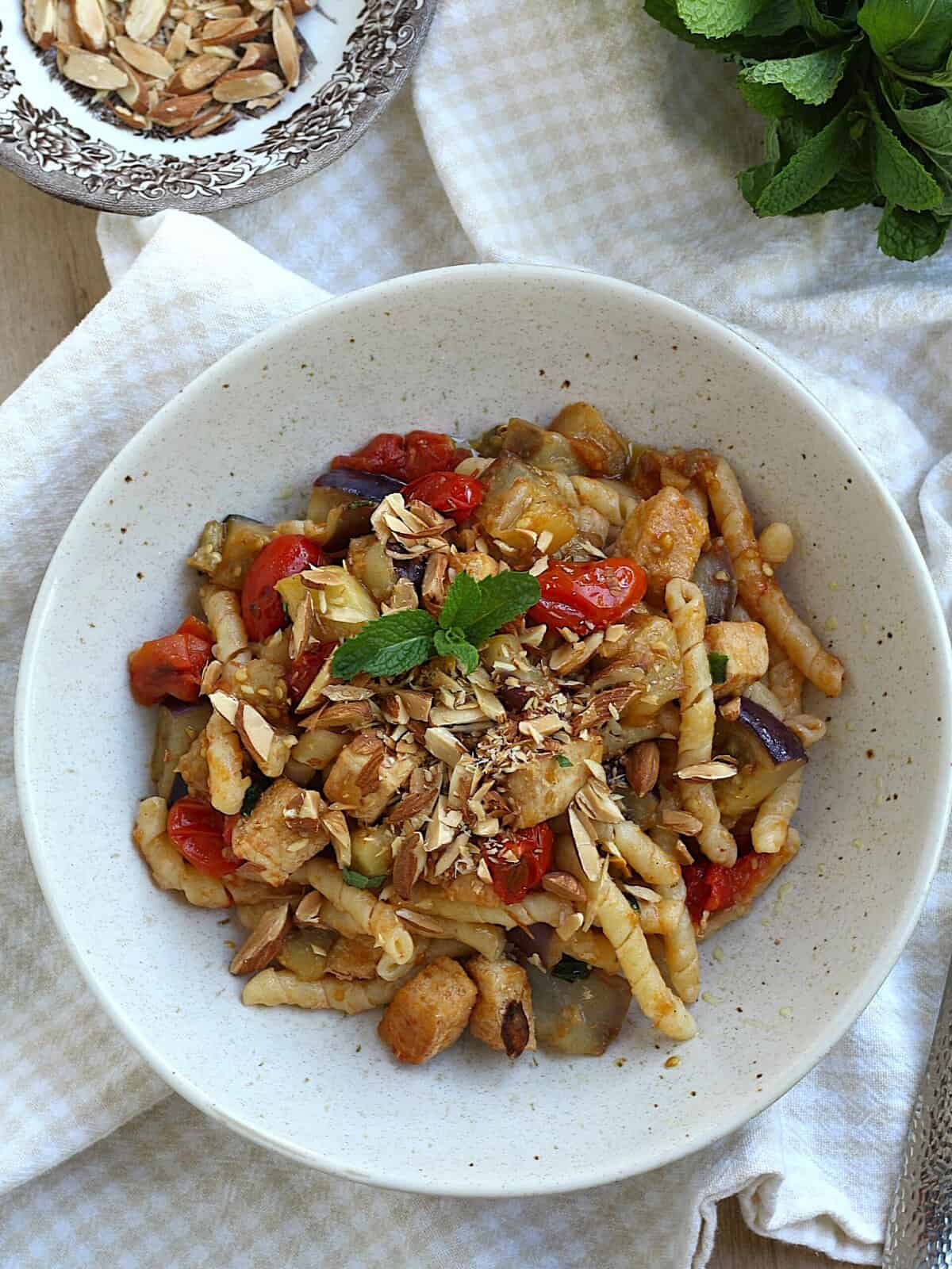 Pasta with eggplant, swordfish and tomatoes topped with mint and chopped almonds in a bowl.