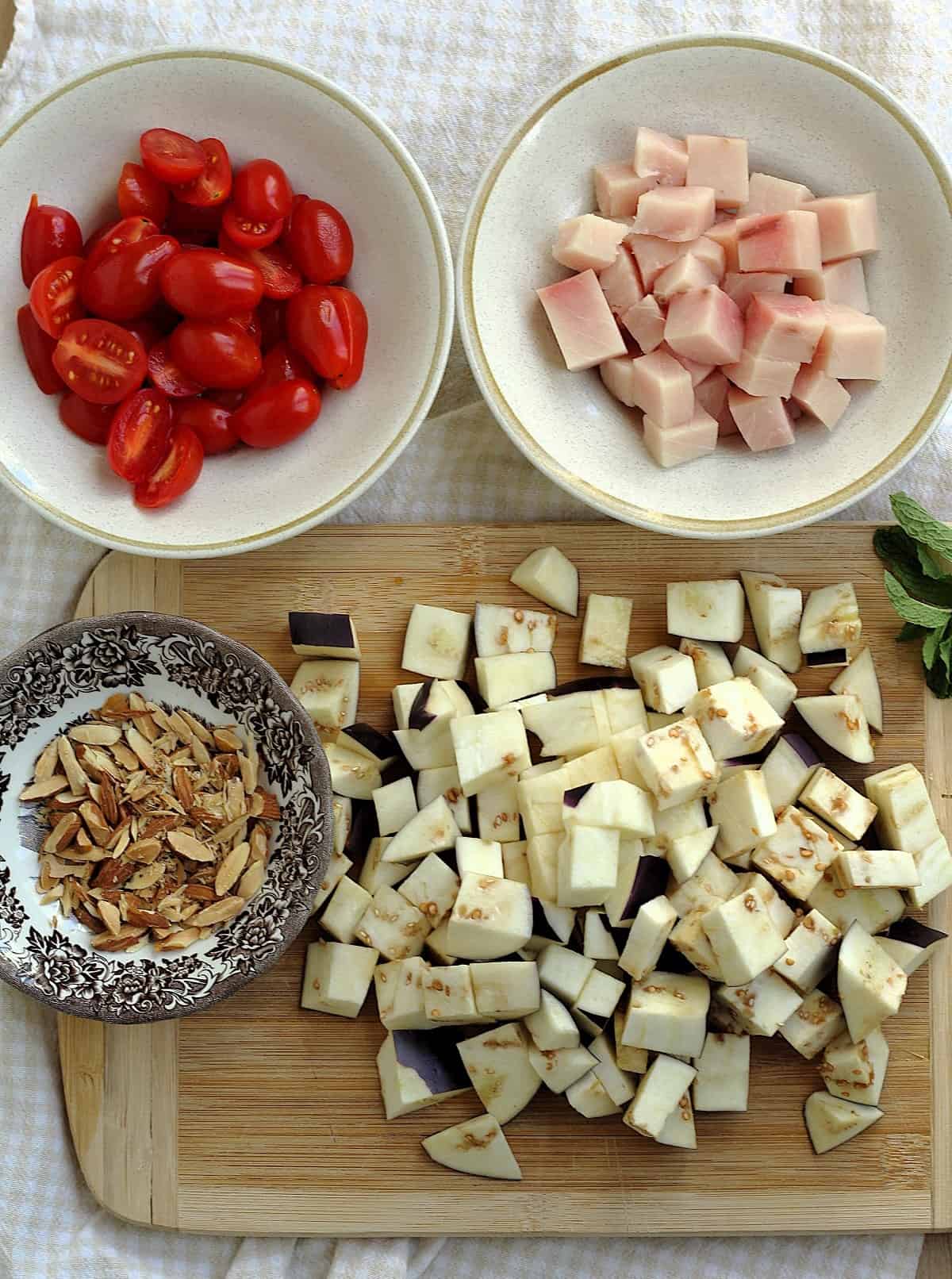 Cubed eggplant, swordfish, halved cherry tomatoes and chopped almonds in bowls.
