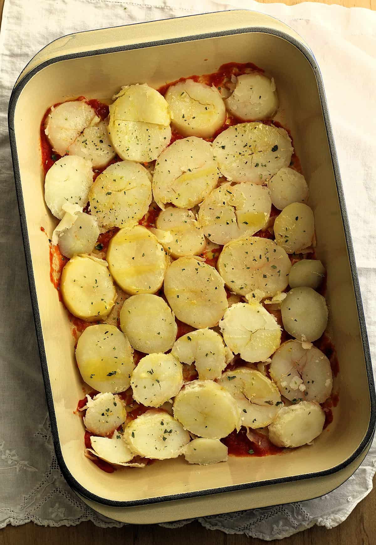 Sliced boiled potatoes layered over tomato sauce in casserole dish.