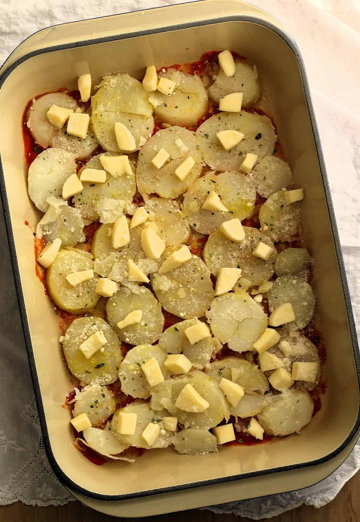 Sliced potatoes topped with oregano and cubed mozzarella cheese in casserole dish.