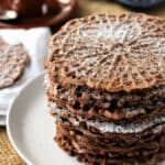 Italian chocolate pizzelle cookies stacked on plate and topped with powdered sugar.