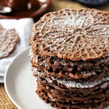 Italian chocolate pizzelle cookies stacked on plate and topped with powdered sugar.