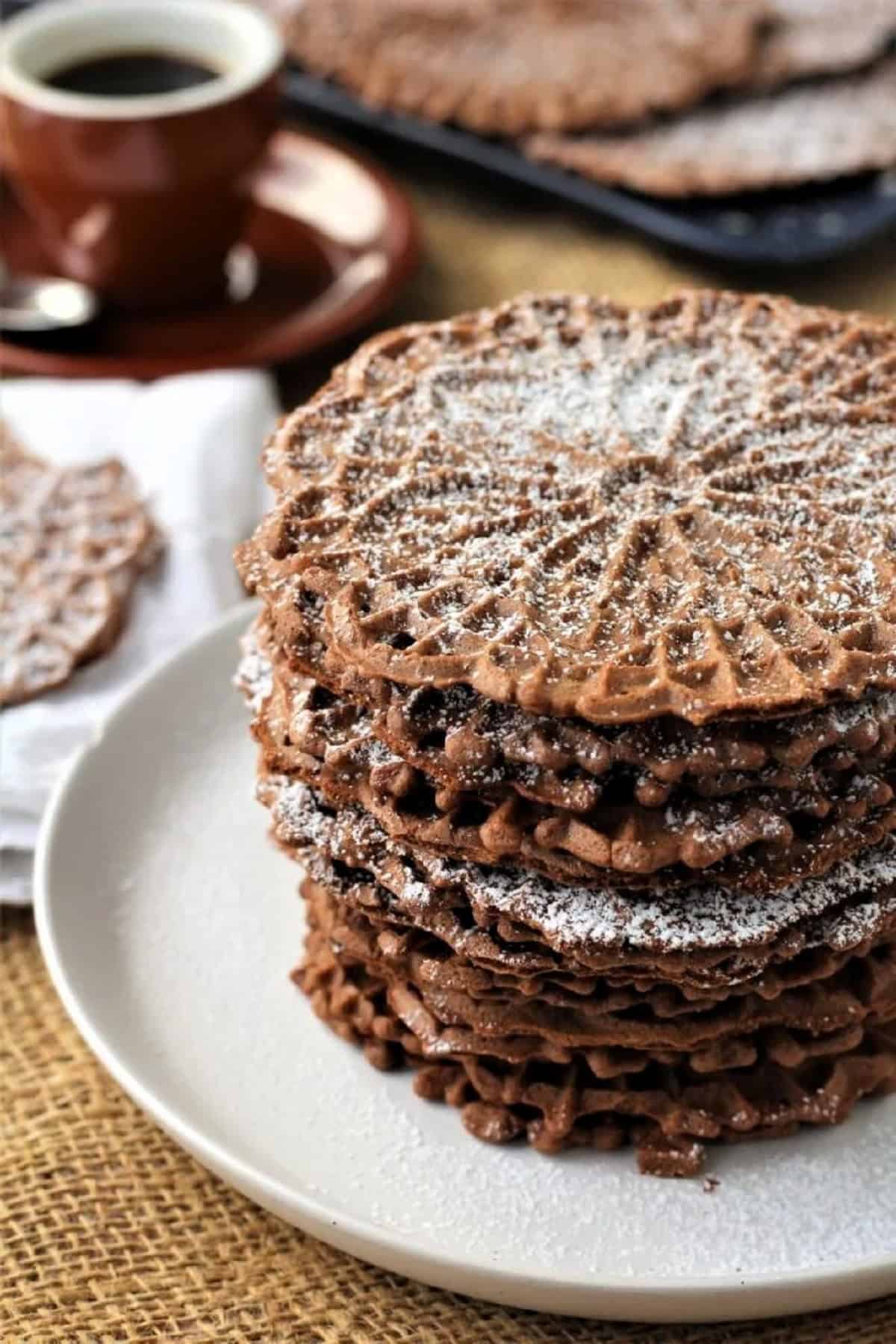 Italian chocolate pizzelle cookies stacked on plate and topped with powdered sugar.