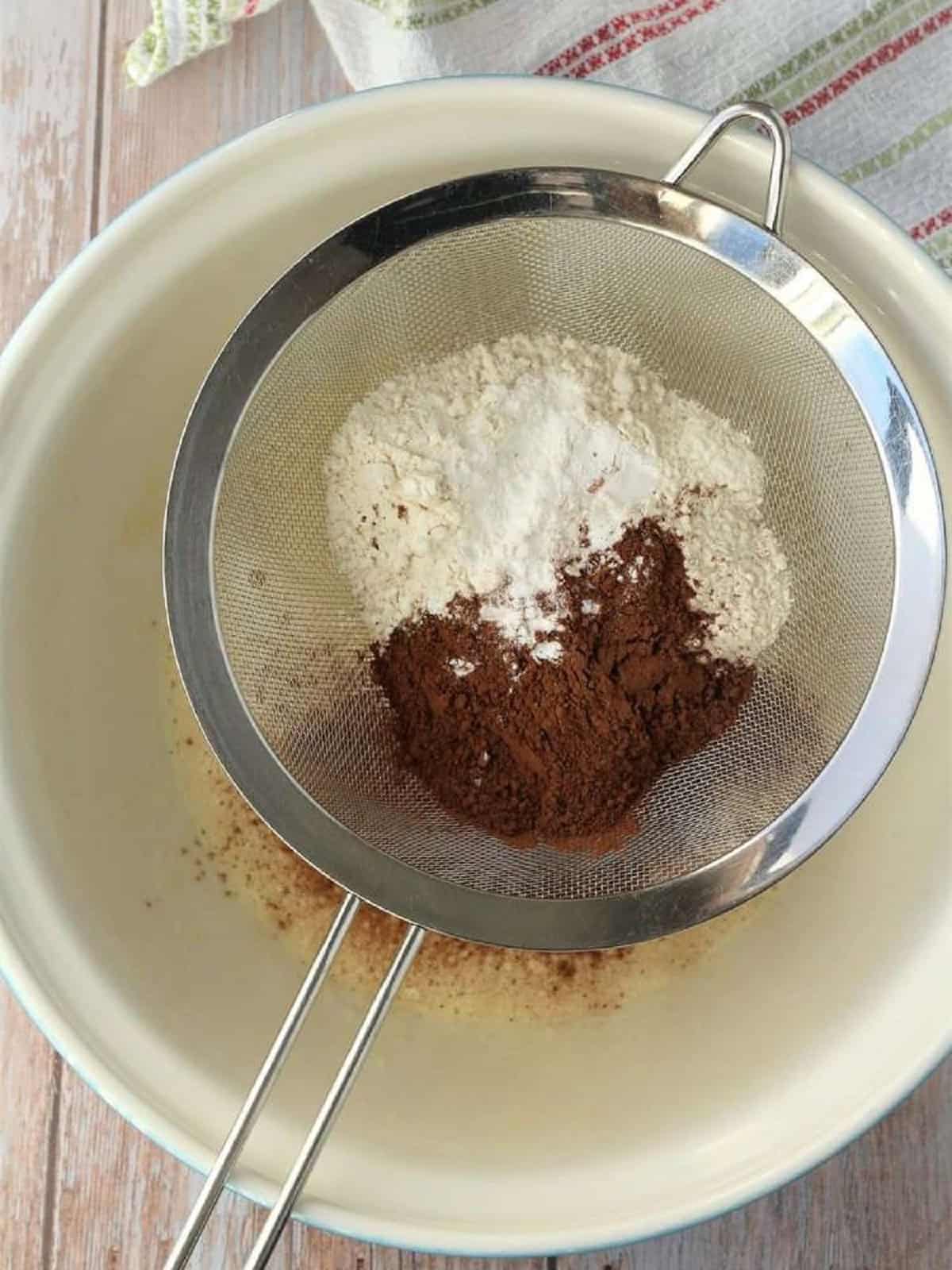 Sifted cocoa powder and baking powder over bowl with wet ingredients for chocolate pizzelle cookies.