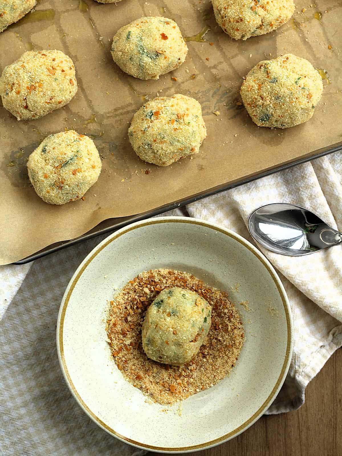 Potato cod croquettes dredged in bowl with breadcrumbs with more patties on baking sheet.