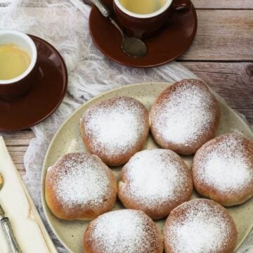 Sicilian iris with ricotta filling topped with powdered sugar on plate next to coffee cups.