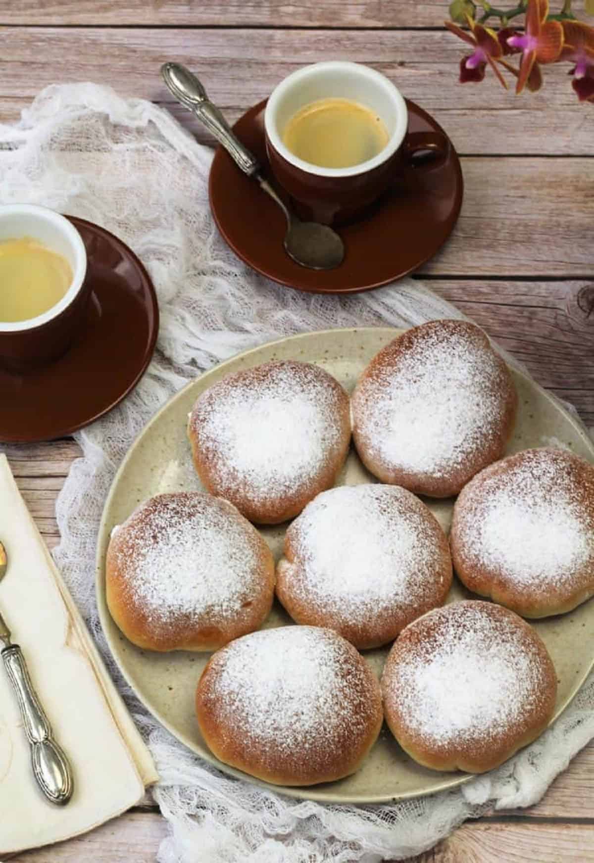 Sicilian iris with ricotta filling topped with powdered sugar on plate next to coffee cups.