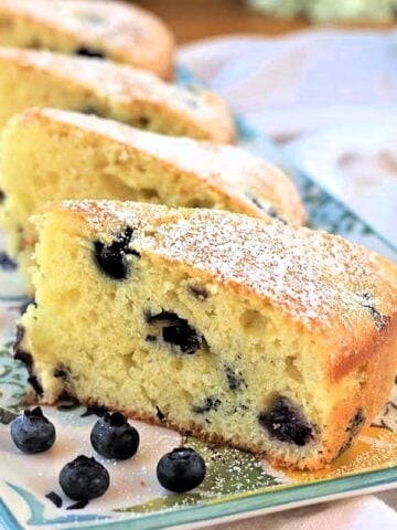 Slices of olive oil cake with blueberries on serving plate with blueberries around it.