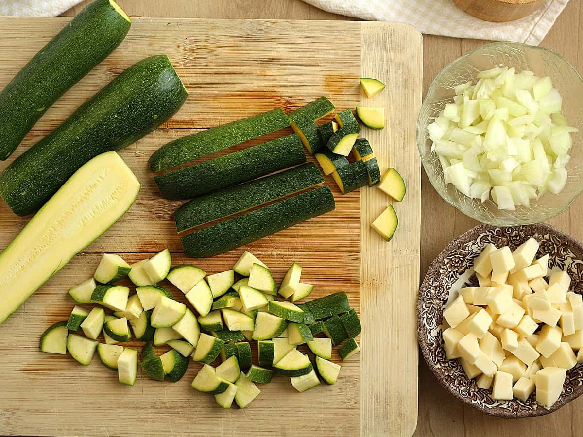 DIced zucchini on wood board, bowl with diced onion and bowl with cubed cheese.