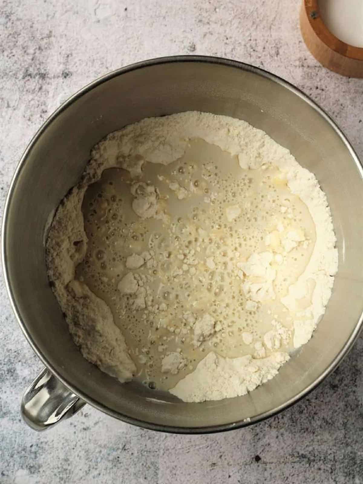 Yeast mixture added to dry ingredients in stand mixer bowl.