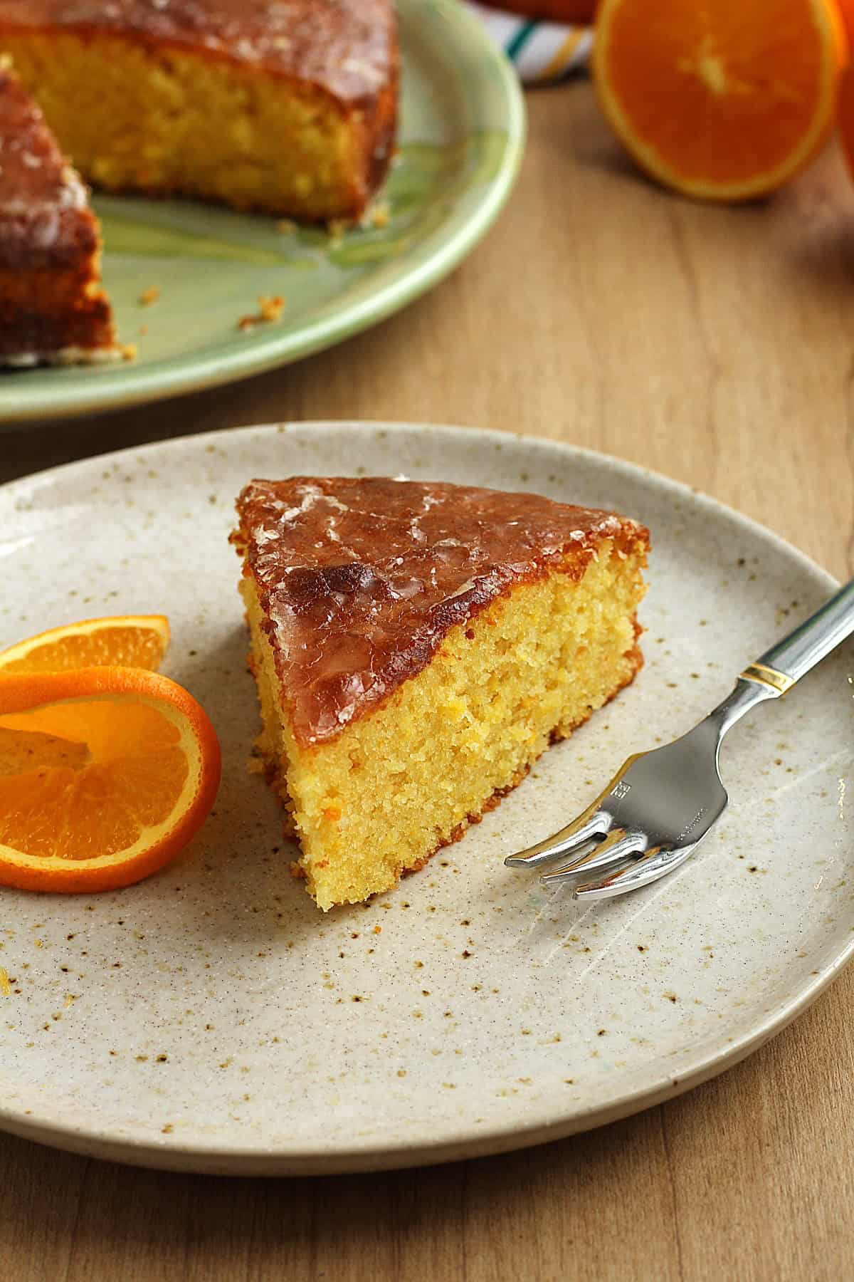 Slice of orange cake with whole orange on plate with fork and orange slice.